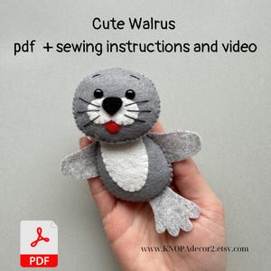 May include: A gray felt walrus plush toy with a white belly and red tongue. The toy is being held in a hand. The text "Cute Walrus" and "pdf + sewing instructions and video" are at the top of the image. A PDF icon is at the bottom of the image with the text "PDF". The text "www.KNOPAdecor2.etsy.com" is at the bottom of the image.