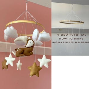 Könnte beinhalten: Ein Baby-Mobile aus Holz mit weißen Wolken, einem braunen Reh und goldenen und weißen Sternen. Das Mobile hängt an einer weißen Decke. Der Text "VIDEO TUTORIAL HOW TO MAKE WOODEN RING FOR BABY MOBILE" ist im Bild sichtbar.