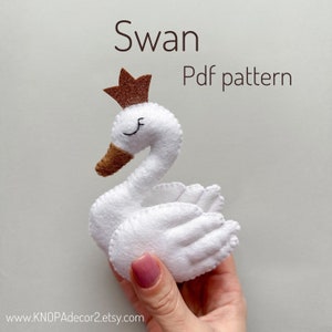 May include: A white felt swan with a brown felt beak and a gold glitter crown. The swan is being held by a hand. The text "Swan Pdf pattern" is written above the swan. The text "www.KNOPAdecor2.etsy.com" is written below the swan.