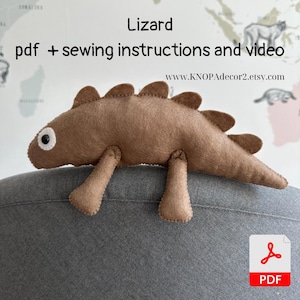以下が含まれることがあります： 白い目をした茶色のフェルトのトカゲが、縫い付けられた笑顔をしています。トカゲは灰色の表面に座っています。画像の上部に「Lizard」というテキストがあり、その下に「pdf + sewing instructions and video」というテキストがあります。画像の下部に「www.KNOPdecor2.etsy.com」というテキストがあります。画像の右下隅にPDFアイコンがあります。