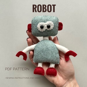 May include: A handcrafted felt robot toy in teal, white, and red. The robot features a rectangular head with large white eyes, red ear details, and red hands and feet. The word "ROBOT" is printed above the toy.