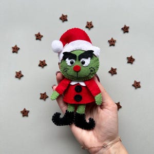 May include: Green felt Christmas ornament of the Grinch wearing a red and white Santa hat. The Grinch has a green face with black eyebrows and a red nose.