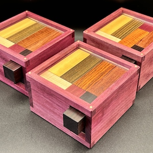 May include: Three wooden puzzle boxes with a vibrant purple exterior and a clear top. The puzzle pieces inside are made of various wood types in shades of yellow, brown, and red. Each box has a dark brown square handle.