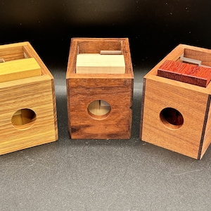 May include: Three wooden puzzle boxes with different colored wood. Each box has a rectangular shape with a circular hole in the front and a sliding top. The boxes contain a light-colored wood block.