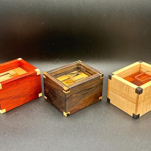 Puede incluir: Tres cajas de rompecabezas de madera en rojo, marrón oscuro y marrón claro. Cada caja tiene forma cuadrada con una parte superior empotrada y detalles en las esquinas. Las cajas se exhiben sobre una superficie oscura.
