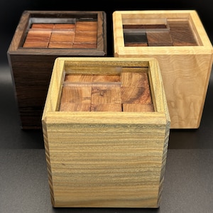 May include: Three wooden puzzle boxes of varying wood tones. Each box is square with a clear plastic insert holding wooden blocks. The boxes are a dark brown, light tan, and a light green-brown.