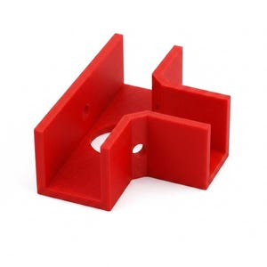 May include: A bright red, L-shaped plastic corner bracket featuring a flat base with two perpendicular walls. The component includes a circular hole in the center of the base and two smaller circular holes positioned on the vertical side walls.