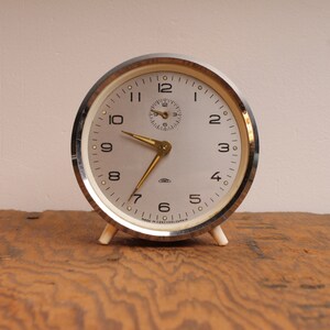 May include: A vintage silver alarm clock with white face and black numbers. The clock has a gold second hand and a small round window for the second hand.