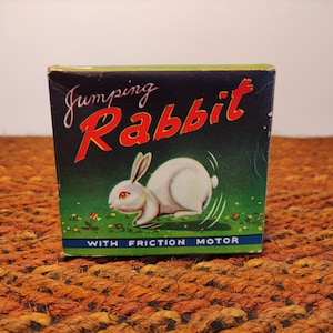 May include: A vintage toy box with a green background and a white rabbit with red eyes jumping. The box says "Jumping Rabbit" in red and black letters and "With Friction Motor" in blue letters.