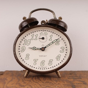 Könnte beinhalten: Ein brauner Vintage-Wecker mit weißem Zifferblatt und schwarzen Zahlen. Die Uhr hat ein kleines Sekundenzeiger-Zifferblatt und das Wort "Peter" ist auf dem Zifferblatt aufgedruckt.