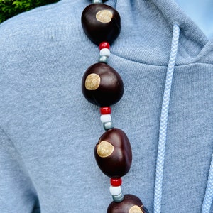 May include: A necklace made of brown buckeyes and white, red, and silver beads. The necklace is worn on a light blue hoodie.