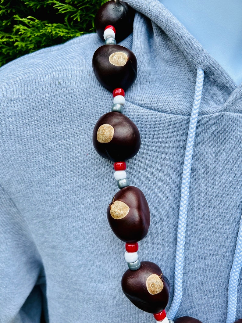 May include: A necklace made of brown buckeyes and white, red, and silver beads. The necklace is worn on a light blue hoodie.