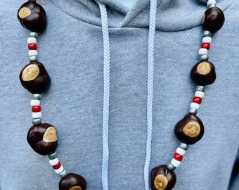 May include: A necklace made of brown buckeyes and white, red, and silver beads. The necklace is worn on a gray hoodie.