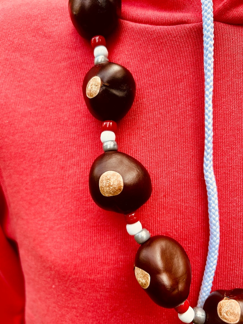 May include: A necklace made of brown, round, natural seed pods with white and red beads. The necklace is worn on a red hoodie.