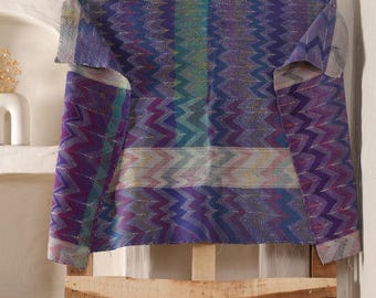 May include: A patterned textile in shades of purple, blue, and green is draped over a wooden pallet. The fabric features a chevron design and has a textured appearance. The textile is folded over the top of the pallet.