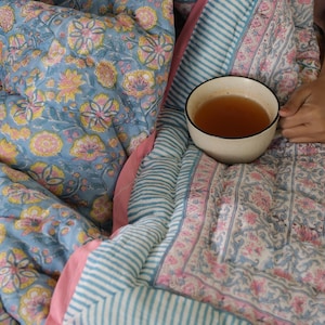 May include: A cozy scene featuring a floral patterned blue quilt with pink and yellow accents. A striped blue and white quilt border and a pink border frame the quilt. A white mug filled with tea is held in the scene.