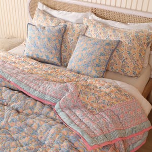 Può includere: Un letto è fatto con una trapunta a motivi floreali, cuscini e federe. La trapunta presenta un motivo floreale blu, rosa e giallo. I cuscini e le federe hanno un motivo simile. La struttura del letto è in legno chiaro.