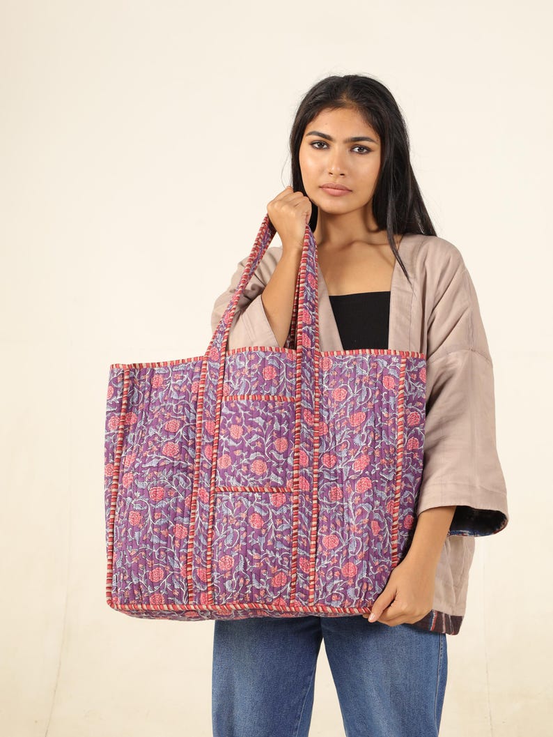 May include: A large purple and pink floral tote bag with a smaller pocket on the front. The bag is made of quilted fabric and has a  stitched design.