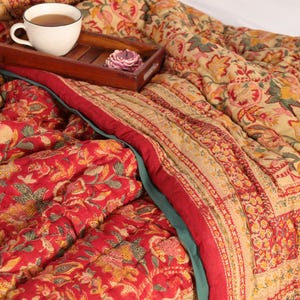 May include: A red and cream floral patterned quilt with a teal border. The quilt is folded and a cup of tea and a tray with a flower are on top of the quilt.