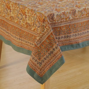 May include: A rectangular tablecloth with a floral pattern in shades of orange, blue, and cream. The border features a repeating design, and the edges are trimmed with a solid teal green. The tablecloth is draped over a table.