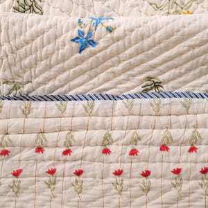 May include: Cream-colored quilted fabric with floral and tree designs. The quilt features rows of red, yellow, and blue flowers, along with green tree and leaf patterns. The fabric has a striped blue and white border.