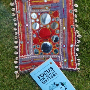 Puede incluir: Un libro azul con el título "Focus on What Matters" de Darius Foroux. El libro está sobre una tela bordada de colores con espejos y conchas.