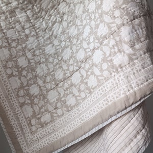 Könnte beinhalten: Eine beige Baumwolldecke mit einem weißen Blumenmuster und einem weißen Rand. Die Decke ist in der Mitte gefaltet und hat auf der Rückseite ein gestreiftes Muster.