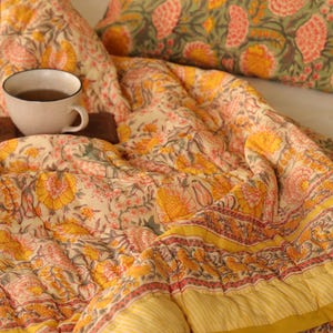 May include: A floral patterned quilt and pillow set in shades of yellow, pink, and green. The quilt features a repeating floral design with a yellow striped border. A cup of tea sits on a wooden tray.