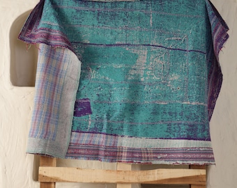 May include: A teal and purple textile with a distressed, textured appearance. The fabric is draped over a wooden structure, revealing a rolled-up section with striped patterns. The edges are finished with a contrasting border, showcasing a vintage or handcrafted aesthetic.