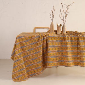 以下が含まれることがあります： オレンジ、グレー、ベージュの水平ストライプの繰り返し模様が施されたマスタードイエローのテーブルクロス。テーブルクロスは、背景に籐の椅子があるテーブルに掛けられています。テーブルには、ドライフラワーの枝が入った装飾的な花瓶が置かれています。