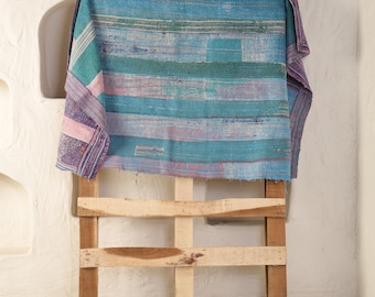 May include: A colorful textile with horizontal stripes in shades of blue, green, and purple is draped over a wooden pallet. The fabric appears to be hand-stitched, with visible seams and variations in color, creating a textured appearance.