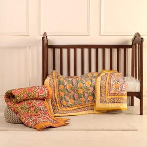 May include: A wooden crib with a floral patterned quilt and matching pillow. The quilt is primarily yellow with pink and green floral designs. A rolled-up quilt rests on a gray ottoman. The crib is made of dark brown wood.