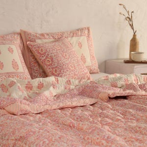 May include: A bed is made with a pink and white floral patterned quilt and matching pillowcases. The quilt and pillowcases have a quilted design. The pillowcases have a white background with a pink floral design.
