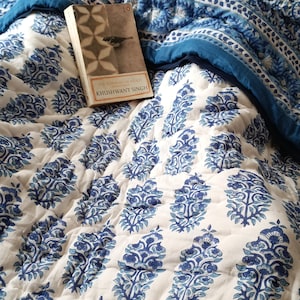 May include: A white and blue patterned quilt with a book titled "The Portrait of a Lady" by Khushwant Singh resting on top.