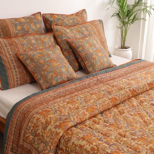 Op de afbeelding: Een beddenset met een bloemenpatroon in oranje, teal en crème. De set bevat een gewatteerde quilt en verschillende bijpassende kussens. De kussens en de quilt hebben een oranje rand. Het beddengoed is op een bed met een wit laken gelegd.