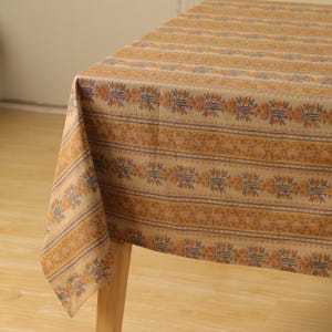 May include: A tan tablecloth with a repeating pattern of floral and striped designs in shades of orange and blue. The tablecloth is draped over a wooden table, showcasing the fabric's texture and design.