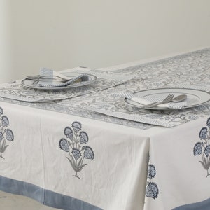 May include: A white tablecloth with a blue floral pattern and a blue border. Two placemats with a blue and white striped pattern are set on the tablecloth. Each placemat has a white plate, a fork, a knife, and a napkin.