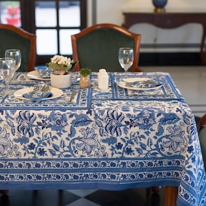 Op de afbeelding: Een wit tafelkleed met een blauw bloemenpatroon en een blauwe rand. Het tafelkleed is gedekt met twee couverts, elk met een wit bord, een blauwe servet en bestek. Er staat een kleine plant in een witte pot op de tafel.