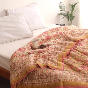 Könnte beinhalten: Eine gesteppte Tagesdecke mit Blumenmuster in Rot-, Rosa- und Gelbtönen, mit einem senfgelben Rand und rosafarbener Einfassung. Die Decke liegt auf einem Bett mit weißen Kissen und weißem Laken, mit einer Tasse auf einem hölzernen Tablett.