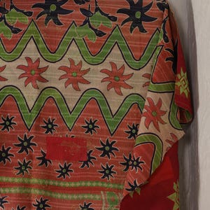 May include: A section of a patterned textile with a red, green, and black floral design. The fabric has a repeating pattern of flowers and zigzags. The textile is folded, revealing a glimpse of the reverse side.