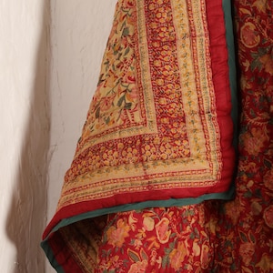 Op de afbeelding: Een rood en crèmekleurig quilt met een bloemenpatroon en een groene rand. Het quilt is dubbelgevouwen en het bloemenpatroon is aan beide kanten zichtbaar.