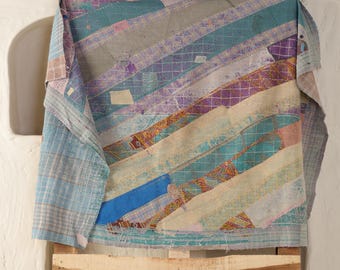 May include: A colorful, patchwork quilt with diagonal stripes in shades of blue, purple, and beige. The quilt is draped over a wooden chair, showcasing its textured surface and intricate design. The handcrafted textile adds a touch of bohemian style.