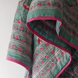 Könnte beinhalten: Eine grüne und rosafarbene Steppdecke mit floralem Muster und rosa Rand. Die Steppdecke ist gefaltet und hängt an einem Haken.