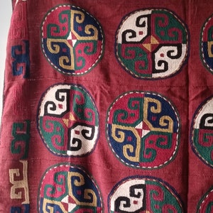 Puede incluir: Un tejido de tela roja con un patrón repetitivo de diseños circulares coloridos. Los diseños están hechos de intrincados remolinos y líneas en tonos de verde, azul, amarillo y blanco.