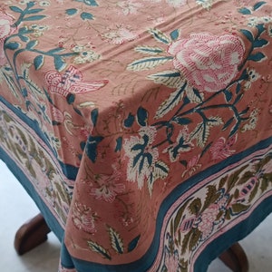 Op de afbeelding: Een bloemig tafelkleed met een perzikachtergrond en een rand van teal en wit. Het tafelkleed heeft grote roze en witte bloemen met teal bladeren en stelen. De rand heeft een herhalend bloemmotief.