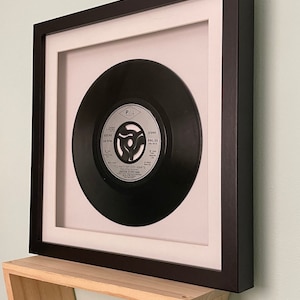 May include: A black framed record display with a white mount and a black vinyl record inside. The record label is silver with the text "PWL Presents" and "Jason Donovan's Too Many Broken Hearts".