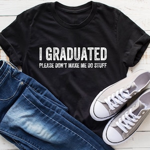 May include: Black t-shirt with white text that reads "I Graduated Please Don't Make Me Do Stuff". The shirt is laid out on a white wooden surface with a pair of blue jeans and gray sneakers.