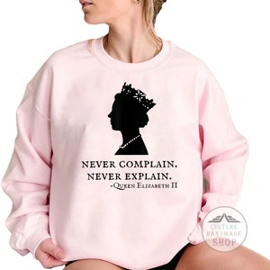 Könnte beinhalten: Rosafarbener Pullover mit einer schwarzen Silhouette von Königin Elisabeth II., die eine Krone und eine Perlenkette trägt. Unter der Silhouette steht der Text "Nie klagen. Nie erklären. -Königin Elisabeth II."