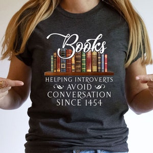 May include: A dark grey t-shirt with a white graphic of a bookshelf full of books. The text on the shirt reads "Books Helping Introverts Avoid Conversation Since 1454."