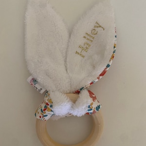 Pode incluir: Um mordedor de coelho branco com um anel de madeira e detalhes em tecido floral. As orelhas de coelho s&atilde;o macias e felpudas, e o anel &eacute; perfeito para as pequenas m&atilde;os agarrarem. O tecido &eacute; estampado com um padr&atilde;o floral colorido. As orelhas de coelho s&atilde;o bordadas com o nome "Hailey".